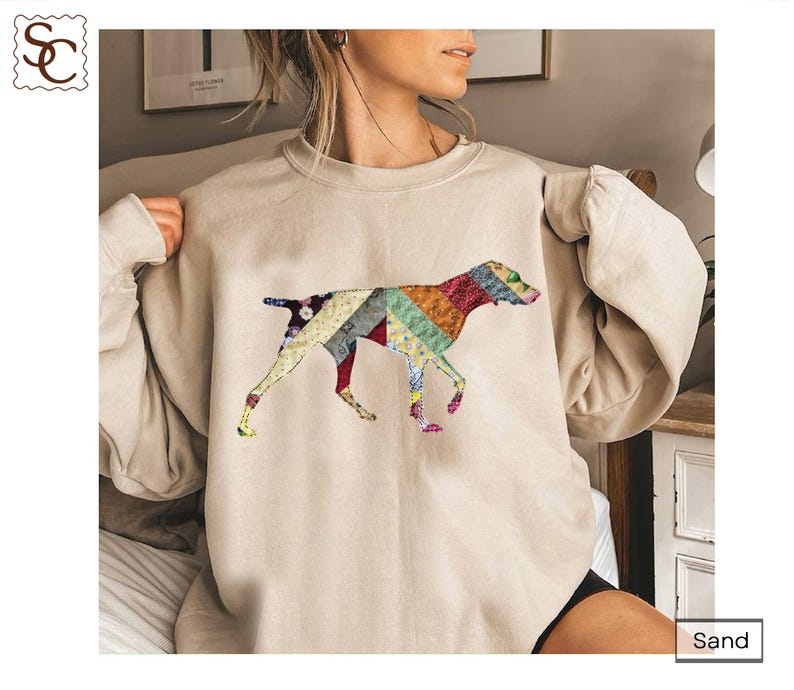 May include: Sand-coloured sweatshirt with a colourful patchwork dog design. The dog is shown walking, with the patchwork design using various colours and patterns. The sweatshirt has long sleeves and a relaxed fit, suitable for casual wear.