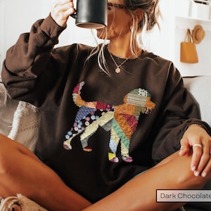 May include: Dark chocolate brown sweatshirt featuring a colorful patchwork dog design. The dog illustration incorporates various fabric patterns and colors, including floral, geometric, and solid hues. The sweatshirt is a relaxed fit and has long sleeves.
