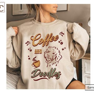 May include: A sand-colored crewneck sweatshirt featuring a graphic print with the text Coffee AND Doodles. The design includes a stylized illustration of a dog face, a steaming coffee cup, and scattered coffee beans in shades of brown and green.