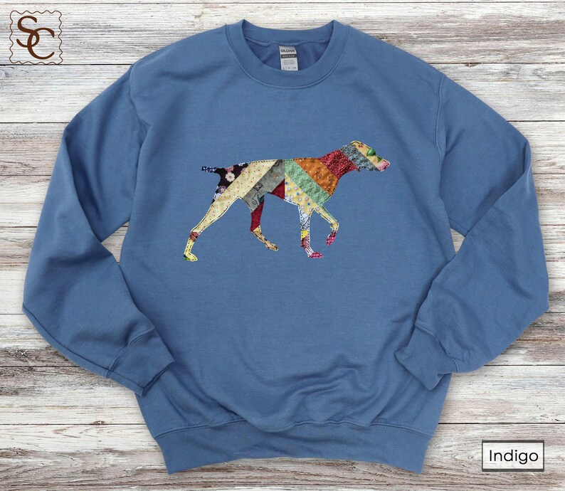 May include: Indigo blue sweatshirt with a colourful patchwork dog design. The dog silhouette is made up of various fabric patterns in shades of red, yellow, green, and blue. The sweatshirt has a crew neck and long sleeves.