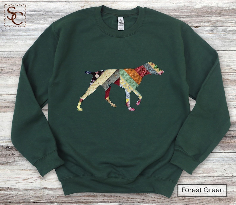 May include: A forest green sweatshirt with a patchwork dog design. The dog is made up of colourful fabric patterns in red, yellow, blue, and green. The sweatshirt has a crew neck and long sleeves. The text "Forest Green" is at the bottom.