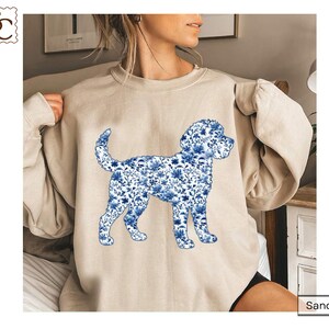 May include: A sand-colored crewneck sweatshirt featuring a graphic of a dog in profile. The dog silhouette is filled with a blue and white floral pattern reminiscent of traditional porcelain designs, set against the solid light beige fabric.