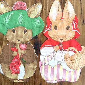 May include: Two fabric cutouts of rabbits. The left rabbit wears a green hat with a pink pom-pom, a brown jacket, and holds a red cloth and a radish. The right rabbit wears a red hood, a white shirt, and carries a woven basket and an umbrella.