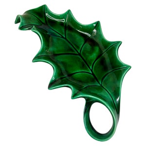 May include: A green ceramic holly leaf dish with a ring handle. The dish is glazed and has a raised vein pattern.