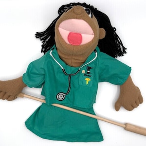 May include: A handmade puppet depicting a doctor, dressed in a teal green scrub top and skirt, with a stethoscope and a "Doctor" patch. The puppet has brown skin, black hair, and an open mouth. A wooden stick is attached.