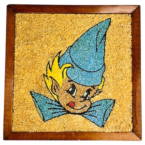 May include: A framed artwork depicting a cartoon elf with a blue hat and bow tie. The artwork is made of small, colorful stones.