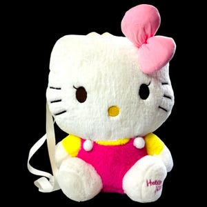 Puede incluir: Una mochila de Hello Kitty de felpa blanca con un gran lazo rosa, ojos marrones y un cuello amarillo. La mochila tiene un mono rosa y correas blancas. Las palabras "Hello Kitty" están bordadas en el pie.