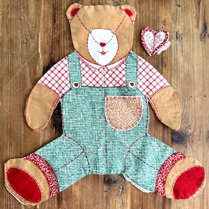 May include: A flat, fabric teddy bear doll featuring a tan head, a red and white checkered shirt, and green overalls with a textured pocket. The bear has red foot pads and a small, separate heart-shaped fabric piece with the text I Love You.