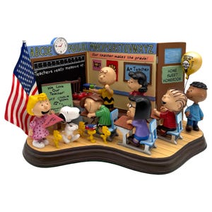 Puede incluir: Una detallada figura que representa una escena escolar con personajes de Peanuts. La escena incluye una bandera estadounidense, un aula con escritorios y una pizarra con texto. Los personajes están en varias poses y se ve un globo.