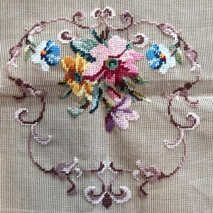 May include: A needlepoint floral design featuring a bouquet of pink, yellow, and purple flowers, with blue accents, enclosed within a brown, ornate border. The background is a beige, grid-like fabric.