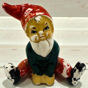 Puede incluir: Una figura de gnomo de jardín de cerámica con un gorro rojo, una camisa verde y pantalones amarillos. El gnomo tiene barba blanca y está sentado con las piernas separadas. La figura tiene zapatos blancos y negros y piernas rojas y blancas.