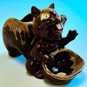 May include: A ceramic squirrel figurine with a brown and tan glaze, holding a bowl with a gold rim. The squirrel has a wide grin, white teeth, and a red mouth. The figurine is a decorative item.