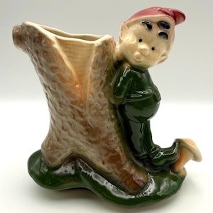 Peut inclure: Un pot de fleurs vintage en céramique représentant un elfe fantaisiste appuyé contre une souche d'arbre. L'elfe porte une tenue verte et un bonnet rouge. Le pot a un intérieur beige et une base verte.