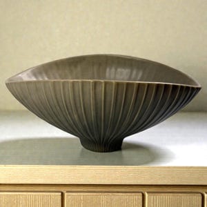 May include: An olive-green ceramic bowl with a fluted exterior design. The bowl has a wide, oval shape and a small, rounded base. The fluted design creates vertical lines that add texture to the bowl's surface. It is likely used for decorative or serving purposes.