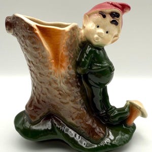 May include: A vintage ceramic planter shaped like a tree trunk with a whimsical elf figure. The elf wears a green outfit and a red hat. The planter has a brown and tan tree trunk with a green base.