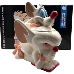 Puede incluir: Un tarro de galletas de cerámica con los personajes de Pinky y Cerebro. El tarro es blanco con detalles en rosa y rojo, incluyendo las orejas, narices y el libro que leen los personajes. La caja detrás del tarro dice "Pinky and the Brain Cookie Jar."