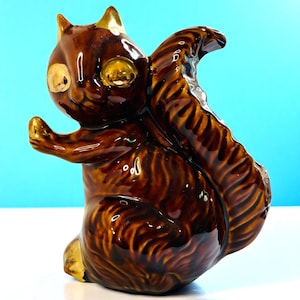May include: A ceramic squirrel figurine in glossy brown glaze with gold accents on the ears, eyes, and paws. The squirrel is posed upright with its tail curved over its back. The figurine has a detailed, textured fur pattern.