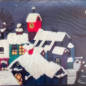 May include: Embroidered winter village scene with snow-covered rooftops, a clock tower, and a full moon. The artwork features a colour palette of blues, whites, greens, and reds, creating a cosy, festive atmosphere. A black cat is on a rooftop.