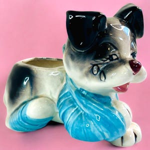 May include: A ceramic planter shaped like a dog with black ears, blue and white fur, and a sad expression. The dog is lying down with its head turned to the side. The planter has a small opening on the top for planting.