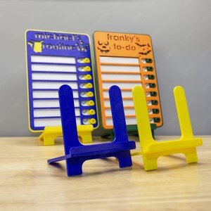 Puede incluir: Dos pizarras de borrado en seco de colores con listas de verificación, una titulada "Rutina de Michael" y la otra "Tareas de Franky", se muestran en caballetes azules y amarillos.