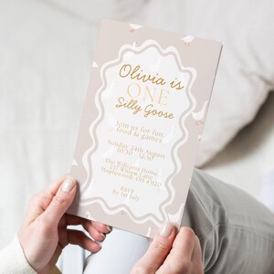 May include: A beige invitation card with gold text and a wavy border. The text reads "Olivia is ONE Silly Goose" with event details. Small goose illustrations are scattered around the border. The card is held by a person.