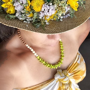 May include: A straw hat adorned with yellow and white flowers and green foliage. A necklace with a light green, spiky design and a section of wooden beads is also visible. The person is wearing a yellow and white patterned top.