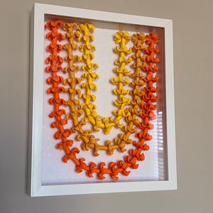 Puede incluir: Una obra de arte enmarcada que presenta una letra "U" estilizada compuesta por elementos florales naranjas, amarillos y dorados. Las flores están dispuestas en un patrón de gradiente, pasando del naranja al amarillo. La obra está encerrada en un marco blanco.