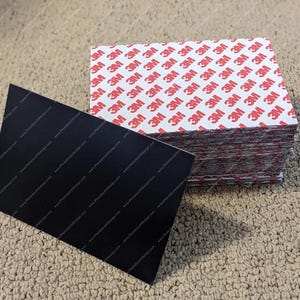 May include: A stack of adhesive squares with the 3M logo printed in red on a white background. A black rectangular piece of material is in the foreground.