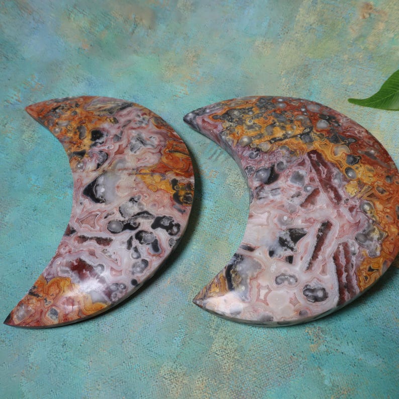 Puede incluir: Dos piedras en forma de media luna con un patr&oacute;n veteado de rojo, naranja, negro y blanco. Las piedras tienen una superficie pulida y se exhiben sobre un fondo turquesa. Estas piedras decorativas son ideales para la decoraci&oacute;n del hogar o como regalos &uacute;nicos.