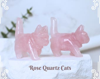 Könnte beinhalten: Zwei rosa Katzenfiguren aus Rosenquarz. Die Katzen sind detailliert geschnitzt und stehen auf einem weißen Steinsockel. Der Text "Rose Quartz Cats" steht darunter.