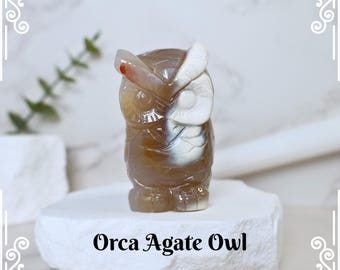 Könnte beinhalten: Orca-Achat-Eulenfigur. Diese geschnitzte Eule weist braune, weiße und rote Farbtöne auf. Die Eule steht auf einer weißen, strukturierten Oberfläche. Der Text "Orca Agate Owl" ist darunter zu sehen.