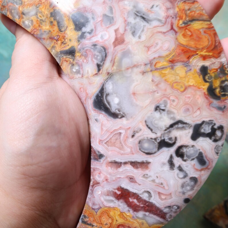 Puede incluir: Una piedra pulida de forma irregular con un patr&oacute;n complejo de colores. La piedra presenta remolinos de rojo, naranja, amarillo, gris y negro, creando un dise&ntilde;o &uacute;nico y natural. La piedra se sostiene en una mano.