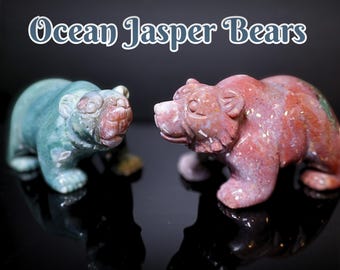 Könnte beinhalten: Zwei Ocean Jasper Bärenfiguren. Ein Bär ist türkis und der andere rot mit weißen Flecken. Die Bären sind mit detaillierten Merkmalen geschnitzt und vor einem schwarzen Hintergrund platziert. Der Text "Ocean Jasper Bears" befindet sich über den Bären.