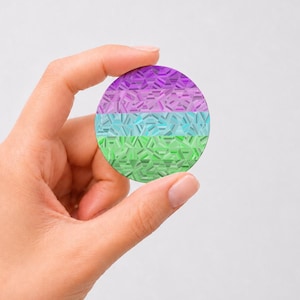 May include: A circular sticker featuring a horizontal tri-color design with purple, light blue, and green bands. The surface has a reflective, holographic finish with a geometric, shattered glass pattern that catches the light across the colors.