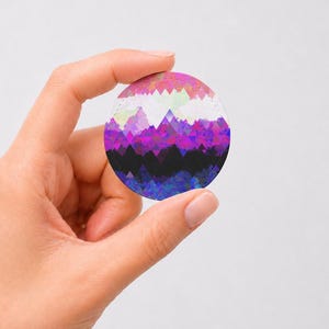 Peut inclure: Un badge circulaire présentant un motif de paysage montagneux géométrique. Le design se compose de triangles empilés dans un dégradé de couleurs, passant du rose clair et blanc au sommet au violet vif, magenta et noir profond à la base.