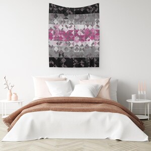 May include: A geometric wall hanging featuring a pattern of triangles in black, grey, and pink. The rectangular tapestry hangs above a bed with white bedding and a brown blanket. The overall theme is modern home decor.