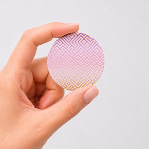 May include: A circular disc featuring a geometric maze pattern in a gradient of yellow, pink, and purple. The design consists of interconnected lines forming a complex path across the entire surface of the round object, which is held between fingers.