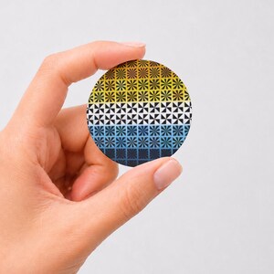 May include: A circular pinback button featuring a repeating geometric starburst pattern. The design transitions in color from a warm yellow at the top, through a white middle section, to a cool blue and dark navy at the bottom edge.