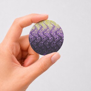 May include: A circular pin featuring a gradient pattern that transitions from a pale yellow at the top to a vibrant purple in the middle and a deep black at the bottom, characterized by a repeating vertical wavy, marbled liquid design.