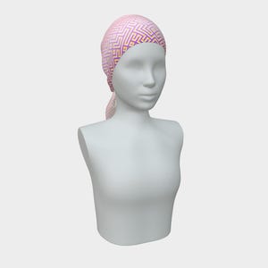 May include: A pink headscarf with a geometric pattern in purple and gold is draped on a mannequin. The head covering is tied at the back of the neck. The mannequin is white and has a neutral expression.
