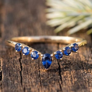 Pode incluir: Um anel de ouro em forma de V com uma safira azul central em corte de pera, ladeada por seis safiras azuis em corte redondo. O aro é de ouro polido e as pedras estão dispostas simetricamente ao longo da borda superior curva do anel.