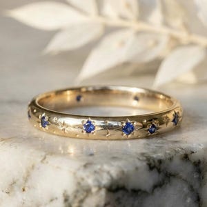 May include: A gold ring featuring small blue gemstones set around the band. The ring is displayed on a marble surface, with star-shaped engravings between the gemstones. A piece of jewellery.
