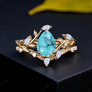 Peut inclure: Une bague en or ornée d'une pierre turquoise en forme de poire et d'accents blancs. La bague présente un design inspiré de la nature avec des détails de feuilles et de branches. La turquoise est d'un bleu-vert vif, sur fond sombre.