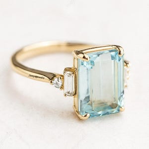 Può includere: Un anello d'oro con una grande pietra preziosa rettangolare azzurra. L'anello ha pietre di accento più piccole e trasparenti su entrambi i lati della pietra principale. L'anello è posto su uno sfondo bianco.