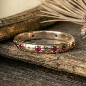 May include: A gold-colored ring with small red gemstones set around the band. The ring has a star and dot pattern engraved between the gemstones. The ring is displayed on a piece of wood with a mirror and pine needles in the background.