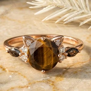 May include: Rose gold ring featuring a large, round tiger's eye gemstone. The ring has a cat-ear design with small diamonds and two dark brown marquise-cut stones on either side of the main stone. The ring is set on a marble surface.