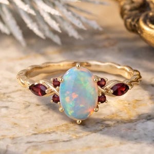 May include: A gold ring featuring a central oval-shaped iridescent opal stone, flanked by small deep red marquise and round-cut garnet gemstones. The ring band has a textured, wavy design, and the setting is crafted with delicate metal prongs.