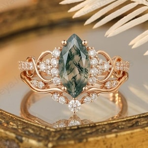 May include: A rose gold ring featuring a marquise-cut green gemstone at its centre, encircled by small, clear gemstones. The ring showcases an intricate design with additional small gemstones along the band. It is displayed on a reflective surface.