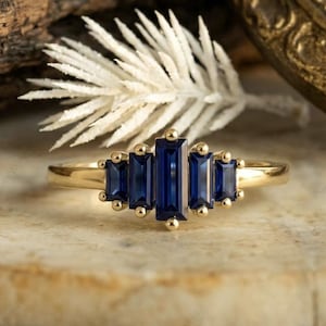 May include: A gold ring featuring five rectangular, deep blue gemstones. The ring's band is a polished gold, and the gemstones are arranged in a linear pattern, with smaller stones flanking a larger central stone. The ring is displayed on a light-colored surface.