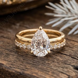 Könnte beinhalten: Ein goldener Verlobungsring-Set mit einem birnenförmigen Diamanten als Mittelstein. Das Set enthält ein Diamantband und ein schlichtes Goldband. Die Ringe werden auf einer rustikalen Holzoberfläche präsentiert.